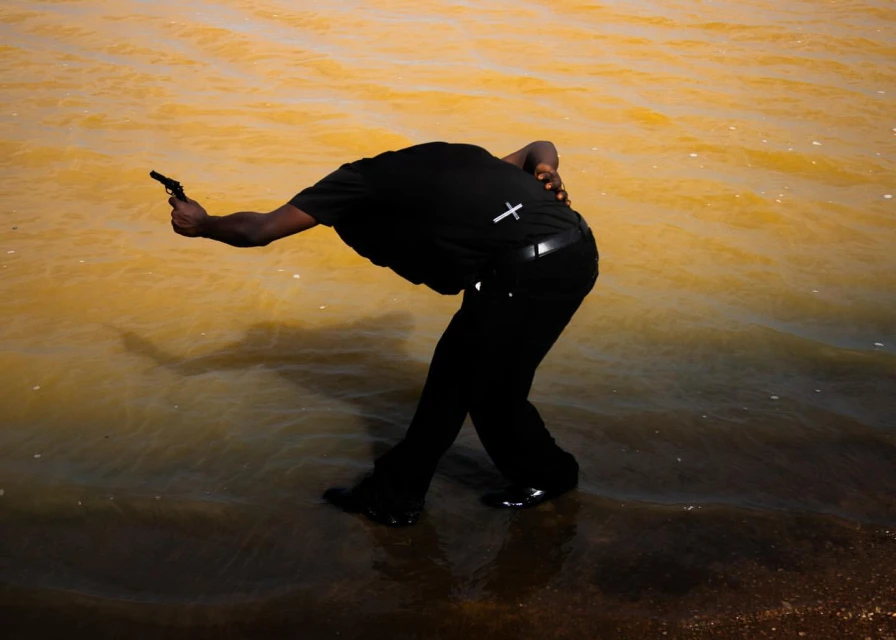 Adult male stands in shallow water with a small handgun, wearing all black with a small cross on his back. 