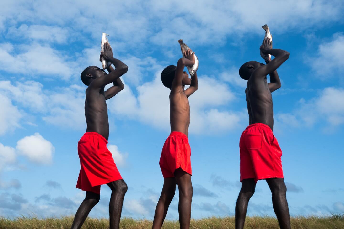 Three bare-chested dark-skinned men in red shorts stand on grassy ground holding fish up to the sky against a partly cloudy blue sky.
