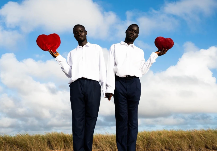 Two dark-skinned men in white shirts and dark pants hold red heart-shaped pillows in a field against a cloudy sky, expressing love.