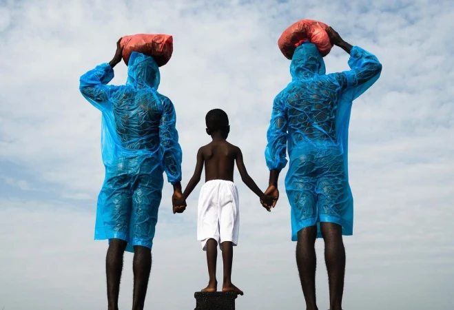 Two adults in blue rain gear hold hands with a child wearing white shorts against a cloudy sky, all standing on a dark surface.