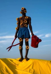 Black man covered in sunflowers wearing blue shorts holding a red watering can against a clear blue sky