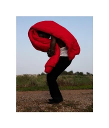 Black man holding a large bright red blanket curved over his upper body outdoors against an open sky.