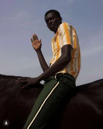Black man wearing a yellow striped shirt and green trousers riding a horse against a pale sky