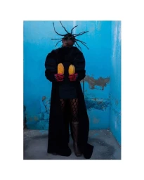 Black model with styled dreadlocks wearing a black coat holding two yellow pods against a textured blue wall.