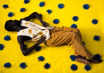 Black man lying on a bright yellow fabric scattered with dark blue artificial flowers in Paris by Nana Yaw Oduro