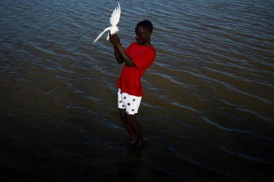 Black man in a red t-shirt and white patterned shorts releasing a white bird into water by photographer Nana Yaw Oduro