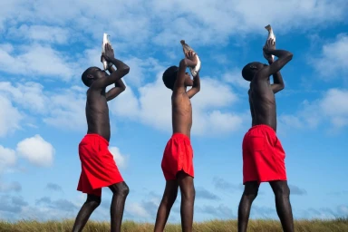 Three shirtless Black men in bright red shorts holding fish over their heads against a bright blue sky and grassy horizon.