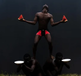 Three black men in a human pyramid formation against a dark background, with the top man holding watermelon slices