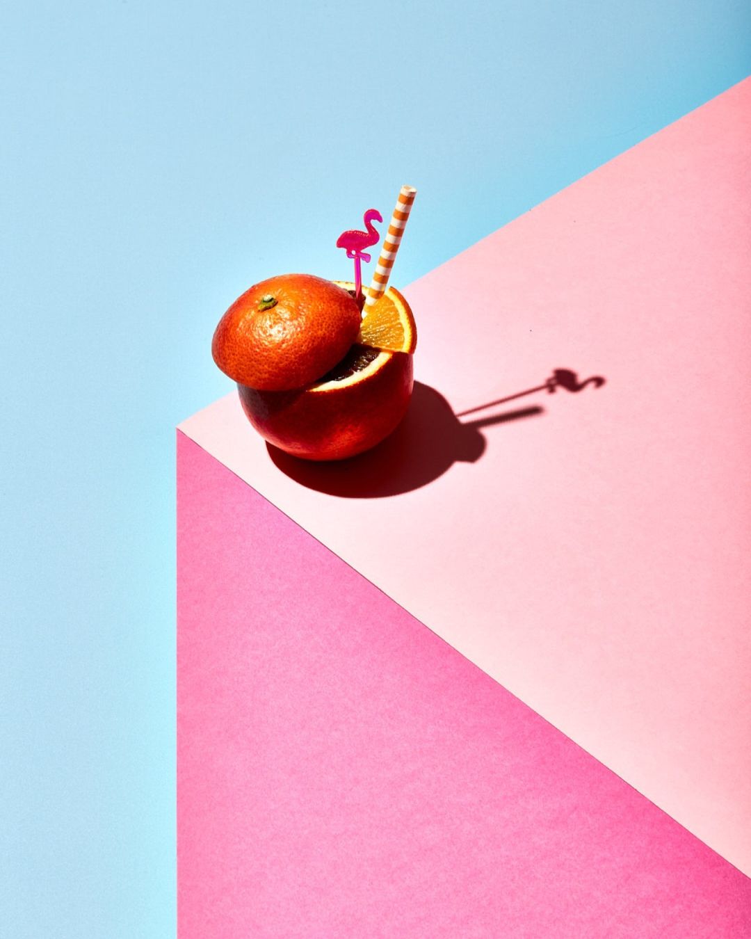 Overhead shot of a halved orange, with a straw and flamingo decoration, serving as a vibrant drink against a geometric backdrop.