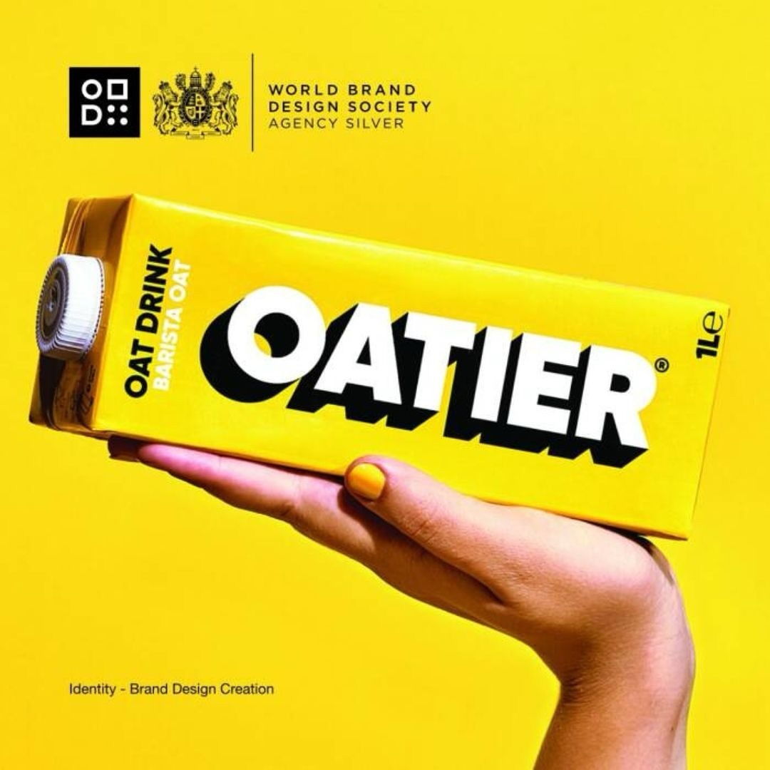 Oatier brand oat milk carton held by a person's hand against a vibrant yellow background, showcasing modern packaging design.