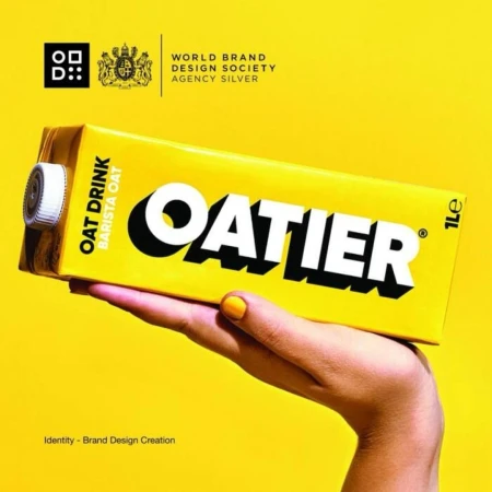 Oatier brand oat milk carton held by a person's hand against a vibrant yellow background, showcasing modern packaging design.