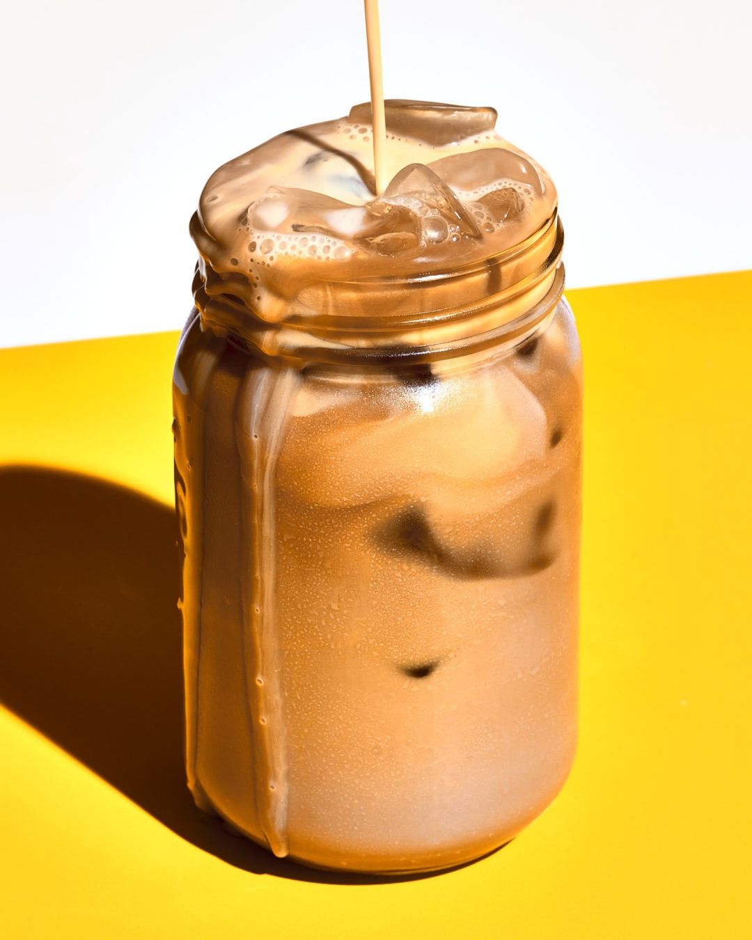 Iced coffee drink with milk being poured into a glass jar with ice, set against a vibrant yellow and white background.