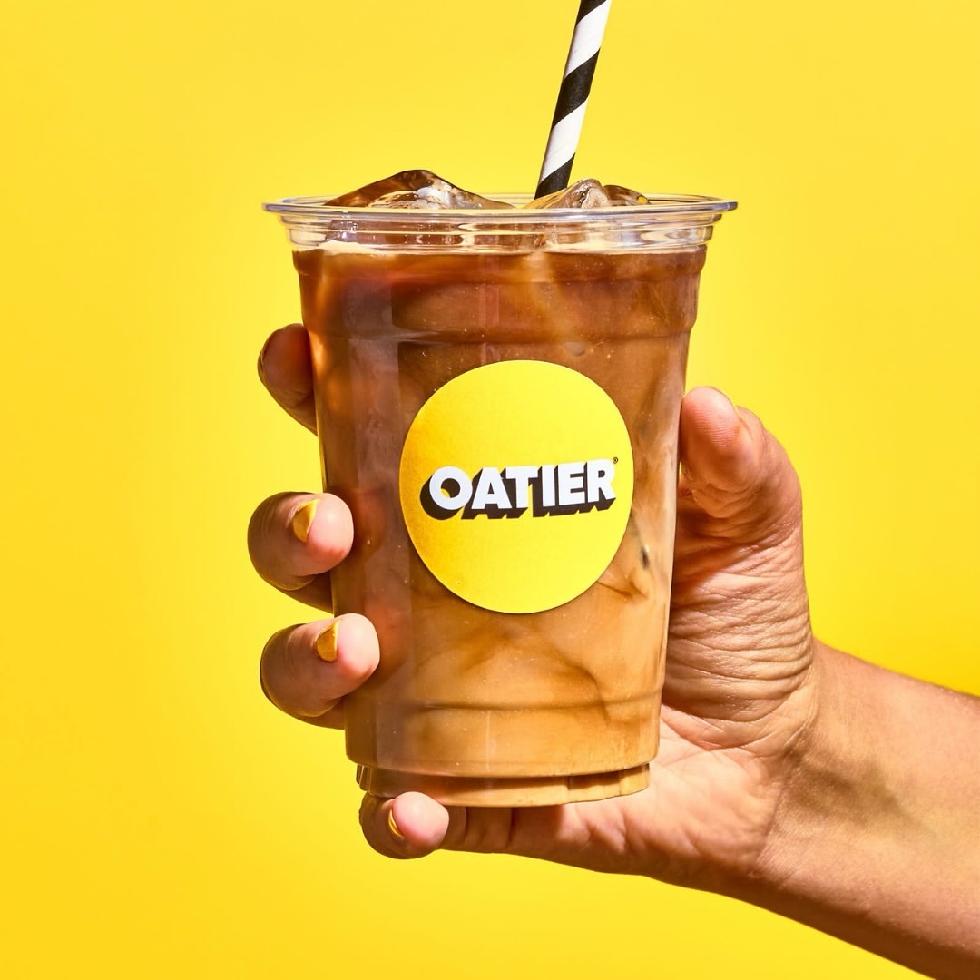 Hand holding iced coffee drink in clear plastic cup with Oatier logo and black-and-white striped straw against a yellow backdrop.