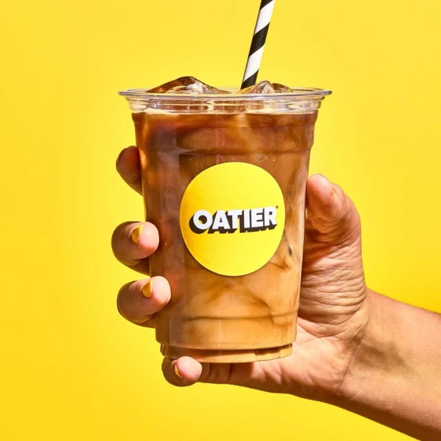 Hand holding iced coffee drink in clear plastic cup with Oatier logo and black-and-white striped straw against a yellow backdrop.