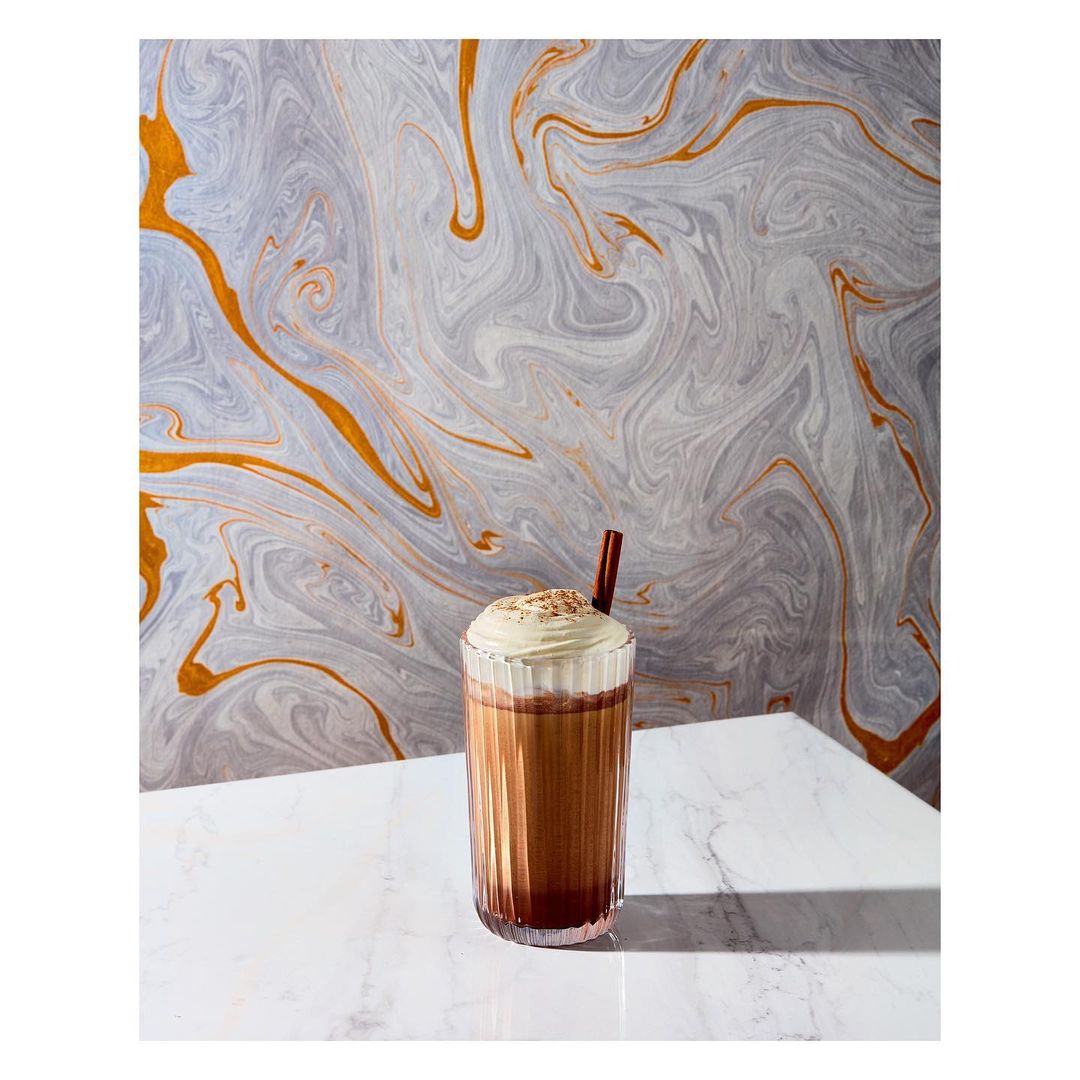Tall glass filled with iced mocha topped with whipped cream and cinnamon stick on white marble table against grey patterned backdrop.