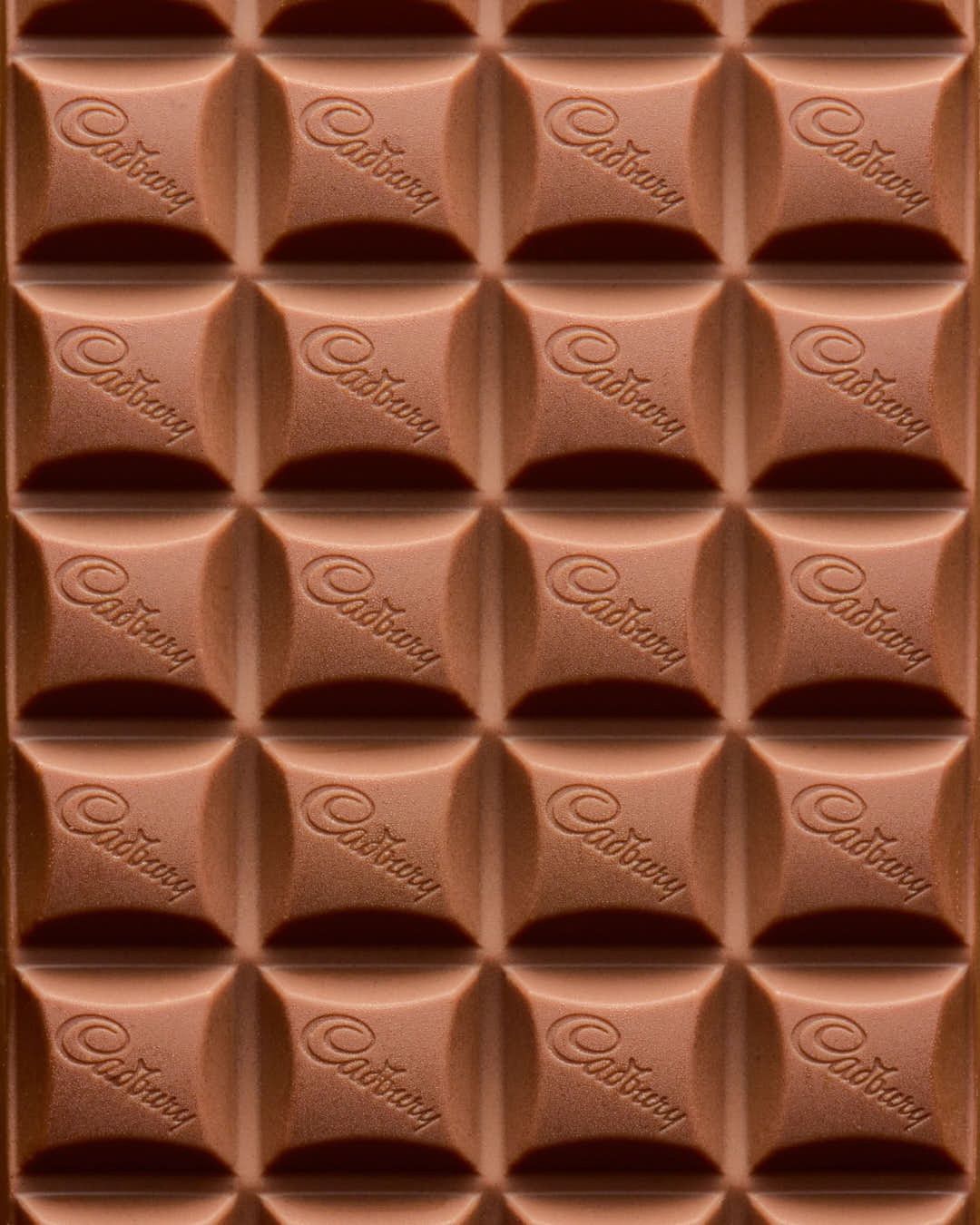 Closeup of a milk chocolate bar with repeating Cadbury logo. The confection is divided into square sections and fills the entire frame.