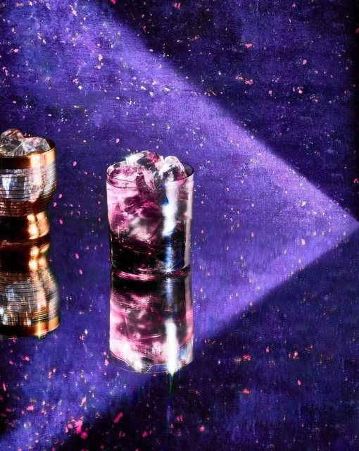 Two cocktail glasses with ice sit on a reflective purple surface against a purple backdrop with scattered flecks of light, bathed in light.