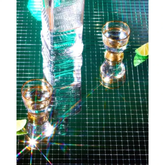Still life shot of a bottle and two glasses on a mirrored surface, featuring clear liquid, limes, and sparkling reflections, creating a vibrant, stylish composition.