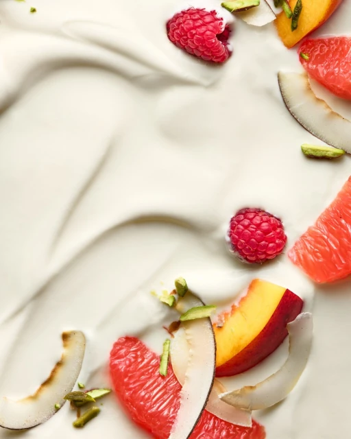 Overhead shot of fresh raspberries, grapefruit, peaches, pistachios and coconut flakes arranged on a swirl of plain white yogurt.