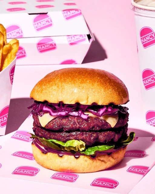 Double cheeseburger with purple cabbage and lettuce, alongside fries and a drink, all branded Nancy on a pink background.