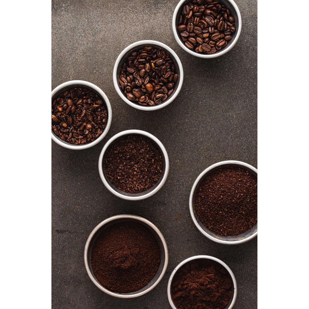 Overhead shot of various types and textures of coffee in white bowls on dark grey surface, including whole beans and ground coffee.