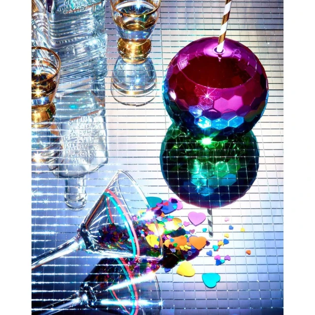 Close-up of a disco ball glass, martini glass with confetti, and glass bottles on a reflective tile surface, creating a vibrant party atmosphere.