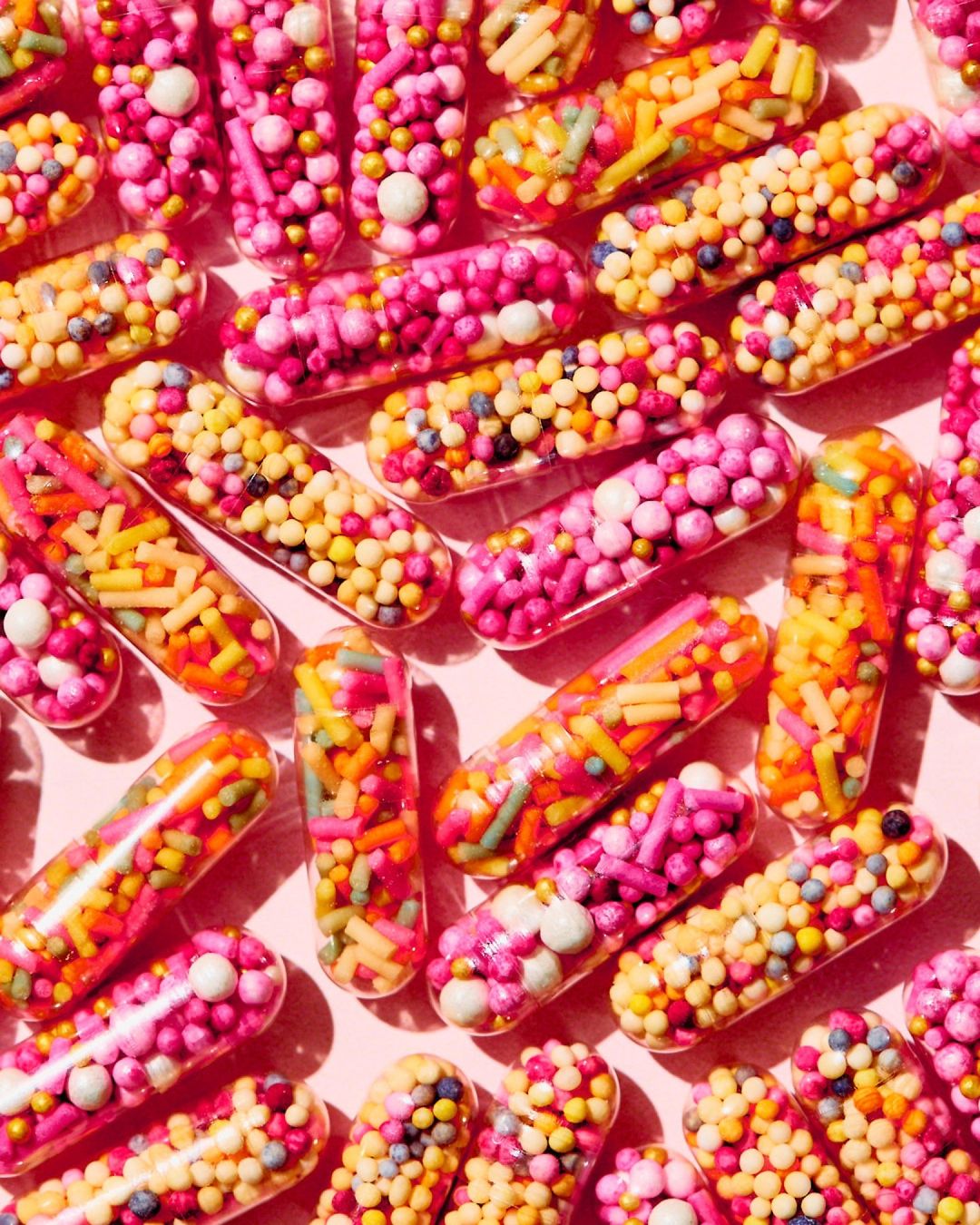 Close-up of numerous clear capsules filled with colorful sprinkles against a light pink background creating a vibrant, abstract pattern.