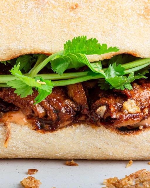 Close-up of a banh mi sandwich featuring pork, cilantro, and sauce on a baguette roll, showcasing its fresh ingredients and textures.