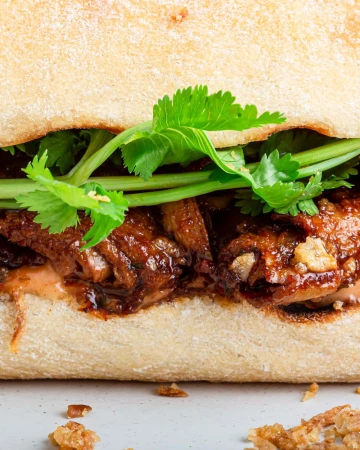 Close-up of a banh mi sandwich featuring pork, cilantro, and sauce on a baguette roll, showcasing its fresh ingredients and textures.