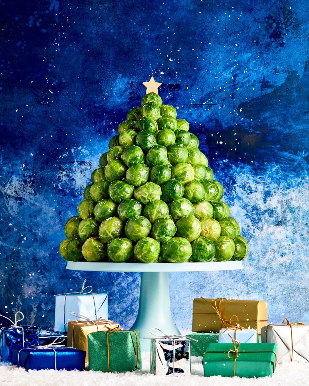 A Christmas tree made of Brussels sprouts on a cake stand surrounded by wrapped gifts against a dark blue backdrop with white spots.