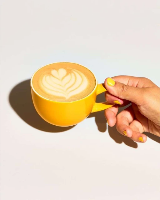 A yellow cup of latte art held in a person's hand with yellow nail polish against a white background in a studio setting.