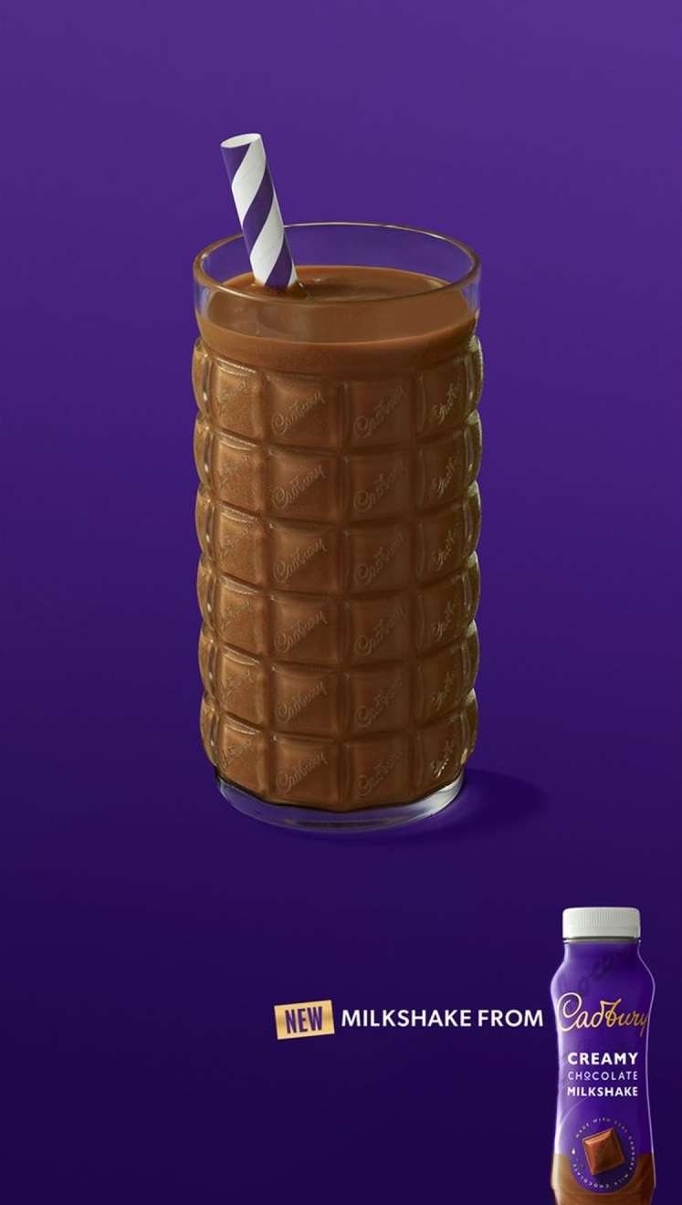 Tall glass filled with chocolate milkshake with striped straw next to bottle of milkshake against purple backdrop in studio lighting.