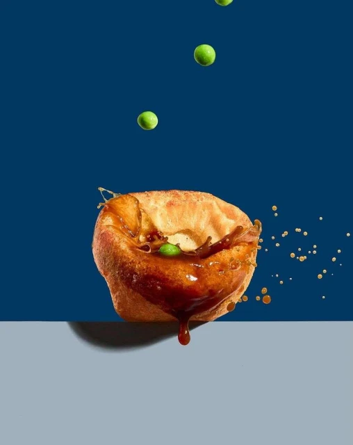 Overhead studio shot of gravy pouring on Yorkshire pudding with green peas in the air against a split blue and grey background.