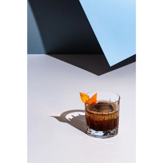 Craft cocktail with ice sphere in glass garnished with orange peel, shadowed on a white surface with gray and light-blue background.