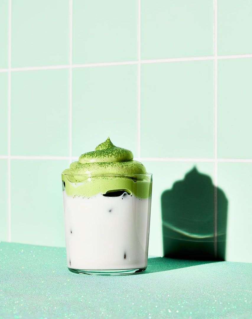 Iced matcha latte with foamy green topping in a clear glass, against a mint green tiled wall and matching table, casting a dark shadow.
