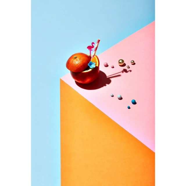 Still life of an orange fruit carved into a cocktail glass with a flamingo straw and surrounded by pink, gold, and blue beads.