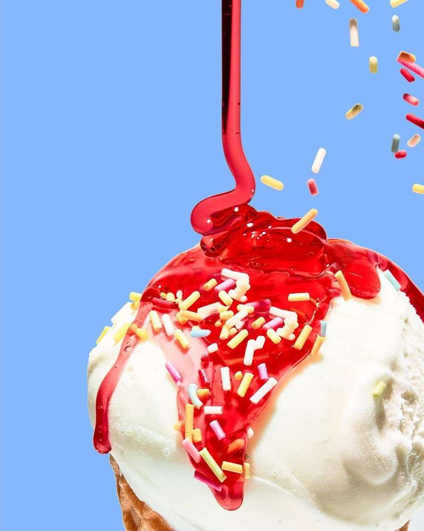 Ice cream cone with white ice cream and a red sauce drizzle with colorful sprinkles, studio shot against a solid light blue backdrop.