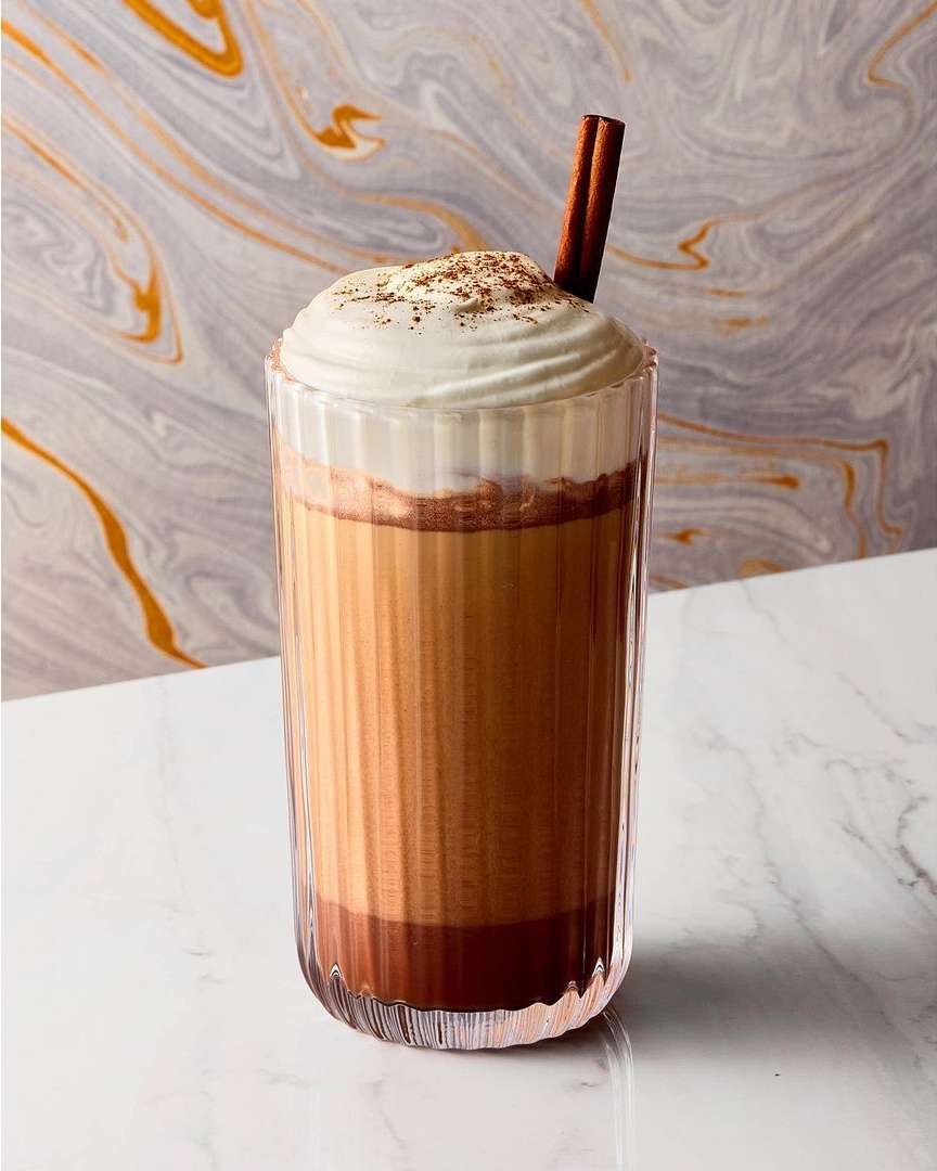A glass of layered beverage topped with whipped cream and a cinnamon stick, placed on a white marble surface against a marble background.