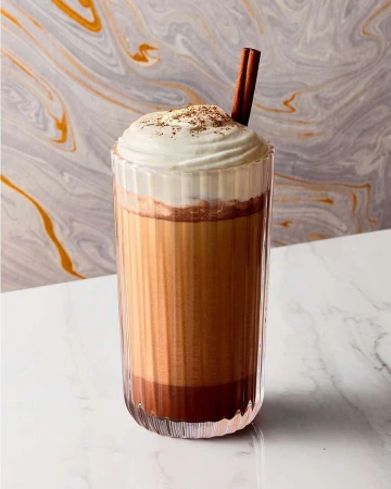 A glass of layered beverage topped with whipped cream and a cinnamon stick, placed on a white marble surface against a marble background.
