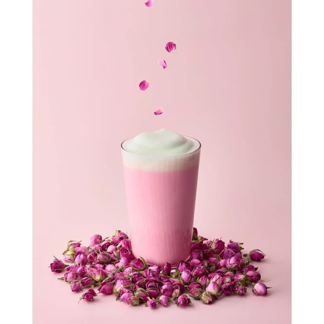 Glass of pink milk with white foam surrounded by dried roses, with falling rose petals against a pink background in studio lighting.