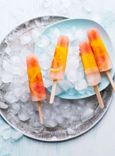 Eye-level close-up of four fruit popsicles on sticks, arranged on ice and a blue plate, atop a silver tray, in bright, natural light.