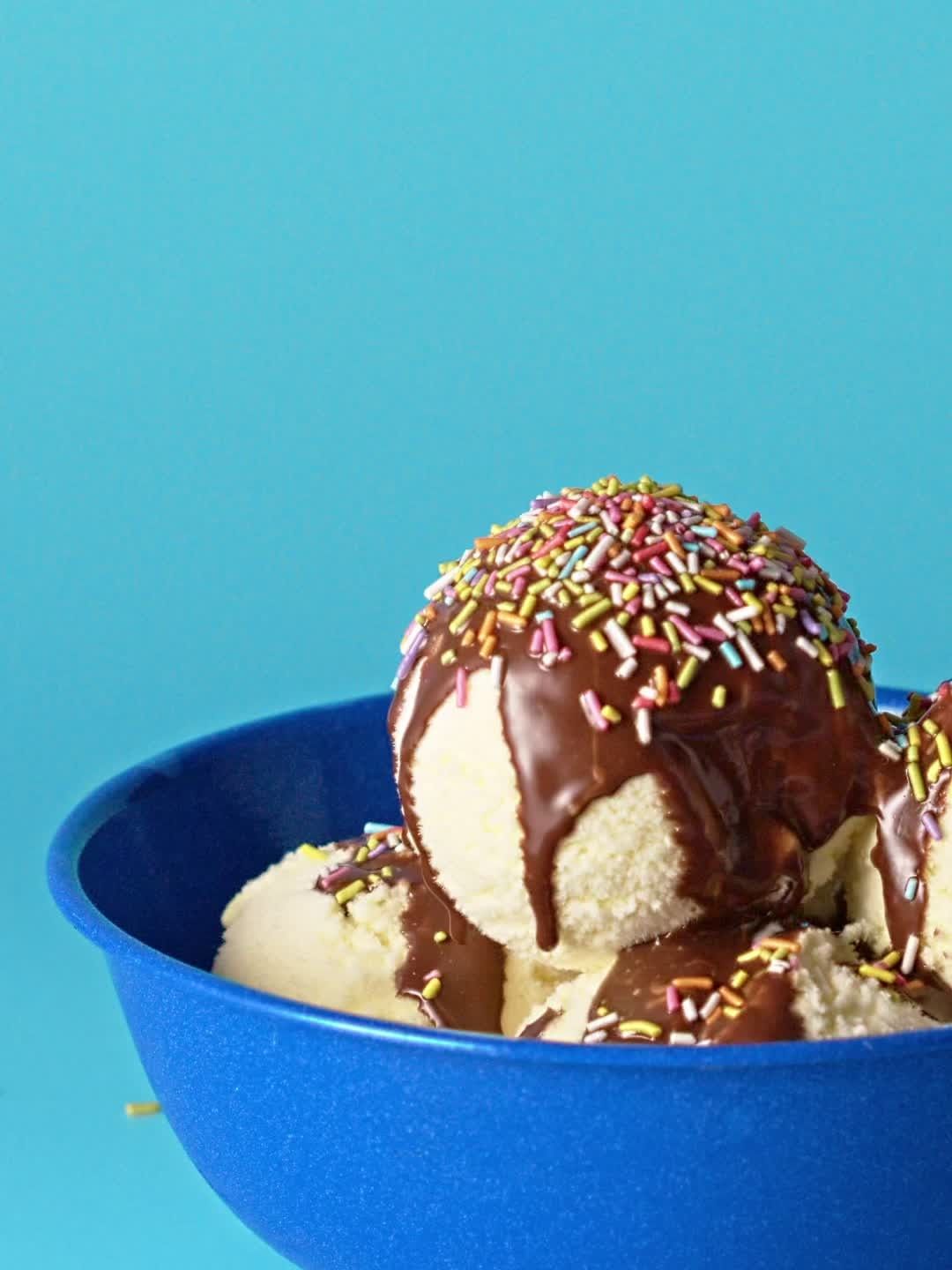 Ice cream scoops in a blue bowl, topped with chocolate syrup and colorful sprinkles against a solid light-blue background.