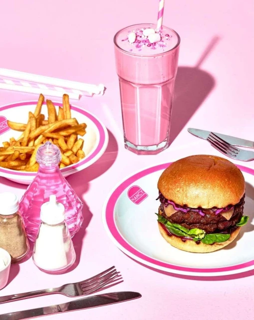 Burger and fries on a white plate with pink trim, a pink milkshake with marshmallows, and condiments on a pink background.