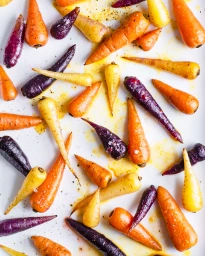 Overhead food photography of roasted orange, purple, and yellow baby carrots scattered on a white surface.