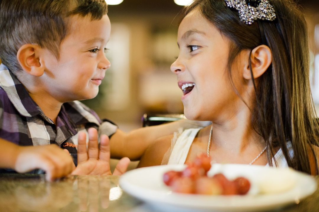 A brother and sister are at a table looking at each other with a plate of grapes and a white substance between them on a light colored table.