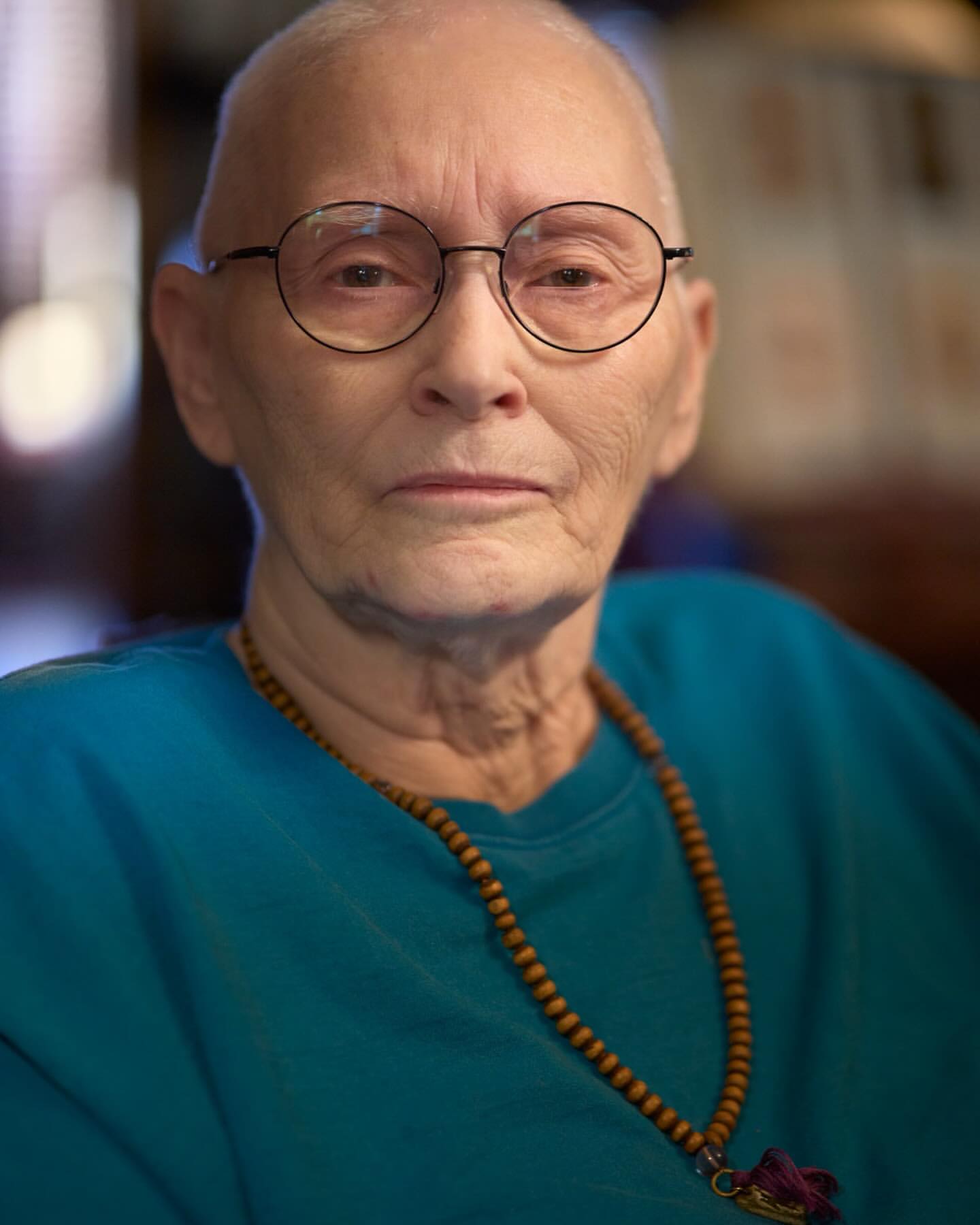 Close-up of a mature woman with bald head, wearing round glasses, a blue shirt, and a beaded necklace looking directly at the camera.