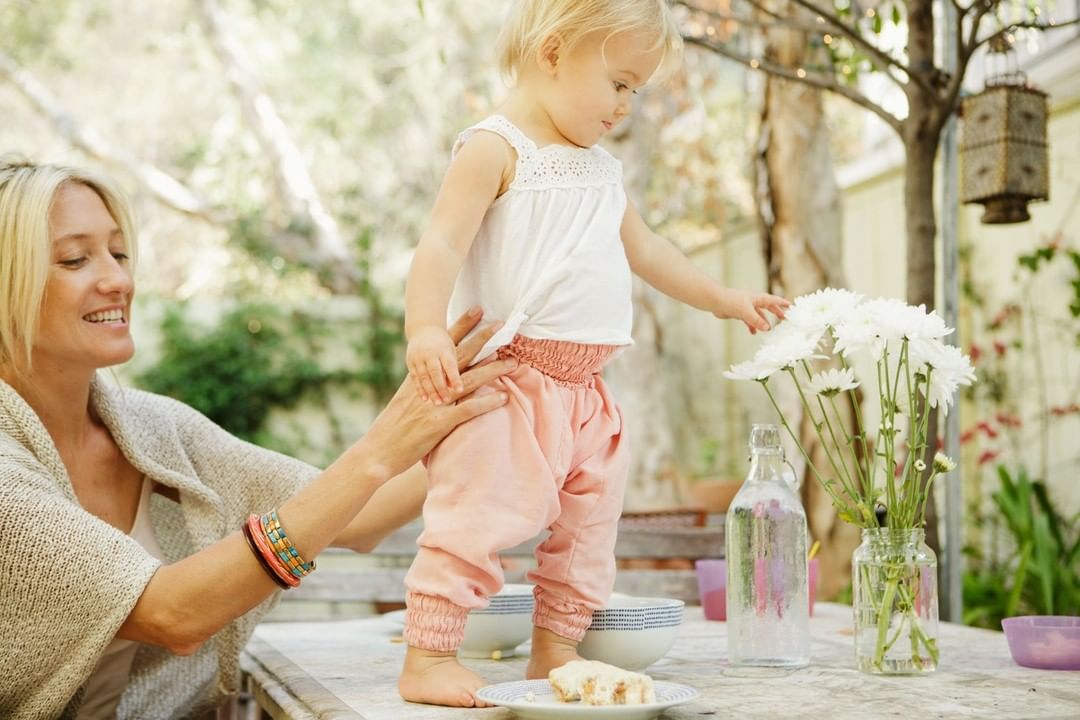 A mother supports her child on a table, with white flowers nearby, in an outdoor setting with dappled light.