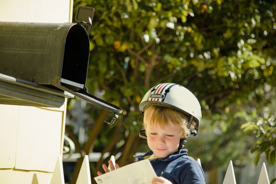 Young boy in helmet reads a letter next to a mailbox, sunny day with lush green foliage, outside with white fence in view.