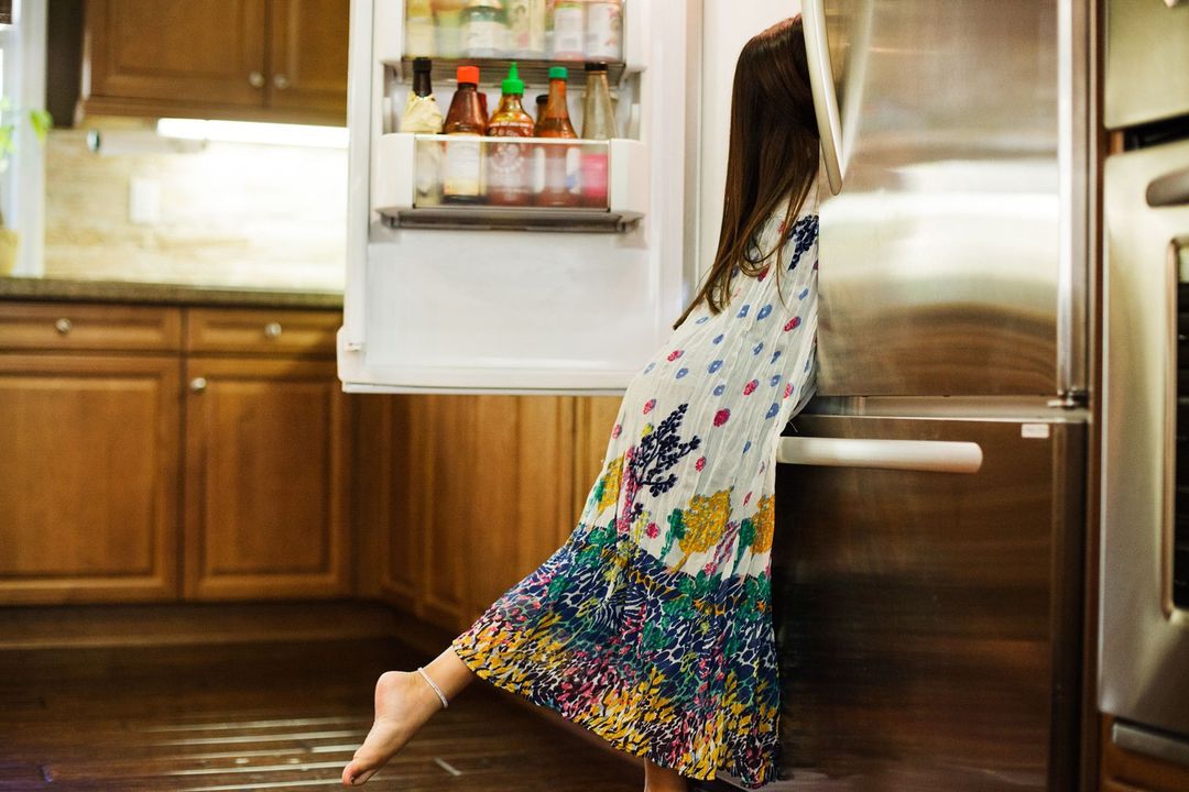 A young girl in a colorful dress leans into an open stainless steel refrigerator in a kitchen to look inside.