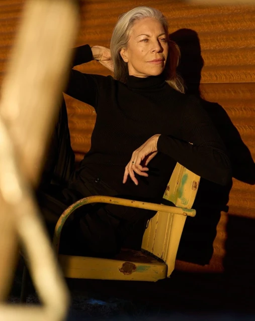 Elegant older woman with gray hair poses in an outdoor portrait, wearing a black turtleneck and sitting in a yellow chair.